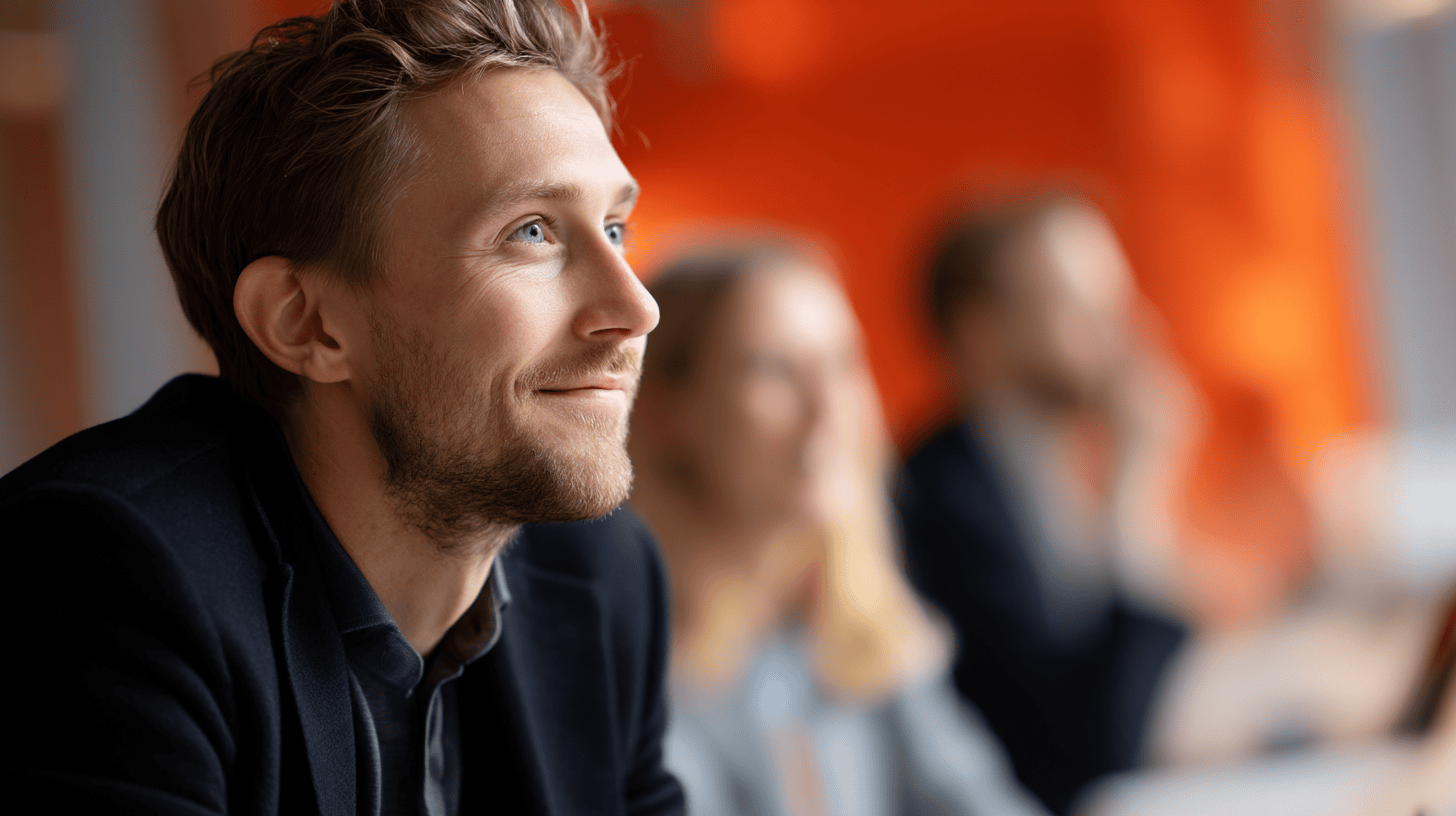 A smiling man with light brown hair and a beard, wearing a dark blazer, looks thoughtfully to the side. In the blurred background, two other people are visible, suggesting a collaborative or thoughtful environment. The setting features an orange wall, indicating a warm atmosphere.