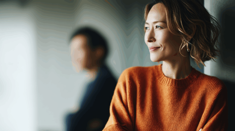 A woman with wavy hair smiles softly, wearing an orange sweater, and stands near a window. A blurred figure of a man is visible in the background, adding a sense of depth to the image. The lighting creates a warm atmosphere, highlighting the woman's expression of contentment and reflection.