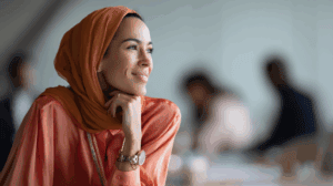 A woman with long dark hair and wearing an orange headscarf sits thoughtfully at a table, resting her chin on her hand and smiling gently. She is dressed in a coral blouse and appears to be in a modern office environment, with blurred figures of other people in the background.