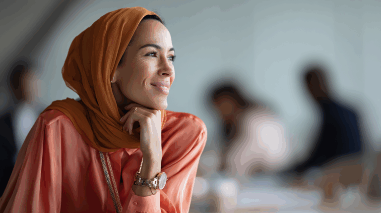 A woman with long dark hair and wearing an orange headscarf sits thoughtfully at a table, resting her chin on her hand and smiling gently. She is dressed in a coral blouse and appears to be in a modern office environment, with blurred figures of other people in the background.