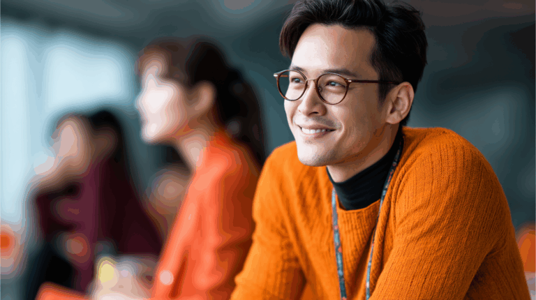 A young man with glasses smiles while sitting at a table. He wears an orange sweater and has a slightly tousled hairstyle. In the background, two blurred figures are visible, one wearing a red garment. The setting appears to be an indoor space with natural light.