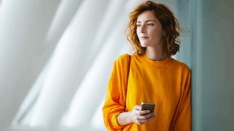 A woman with curly hair is standing indoors, leaning against a wall. She wears a bright orange long-sleeve top and holds a smartphone in one hand while looking thoughtfully into the distance. Soft, natural light filters through large windows, creating a serene atmosphere.
