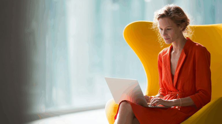 A woman sits comfortably in a bright yellow chair, focused on her laptop. She wears a vibrant orange dress and has wavy hair. The background features soft natural light, creating a serene workspace atmosphere.