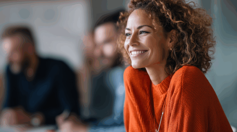 A smiling woman with curly hair wearing an orange sweater sits in a meeting, looking pleasantly engaged. Blurred individuals are visible in the background, suggesting a collaborative environment. The setting is bright and modern, reflecting a professional atmosphere.
