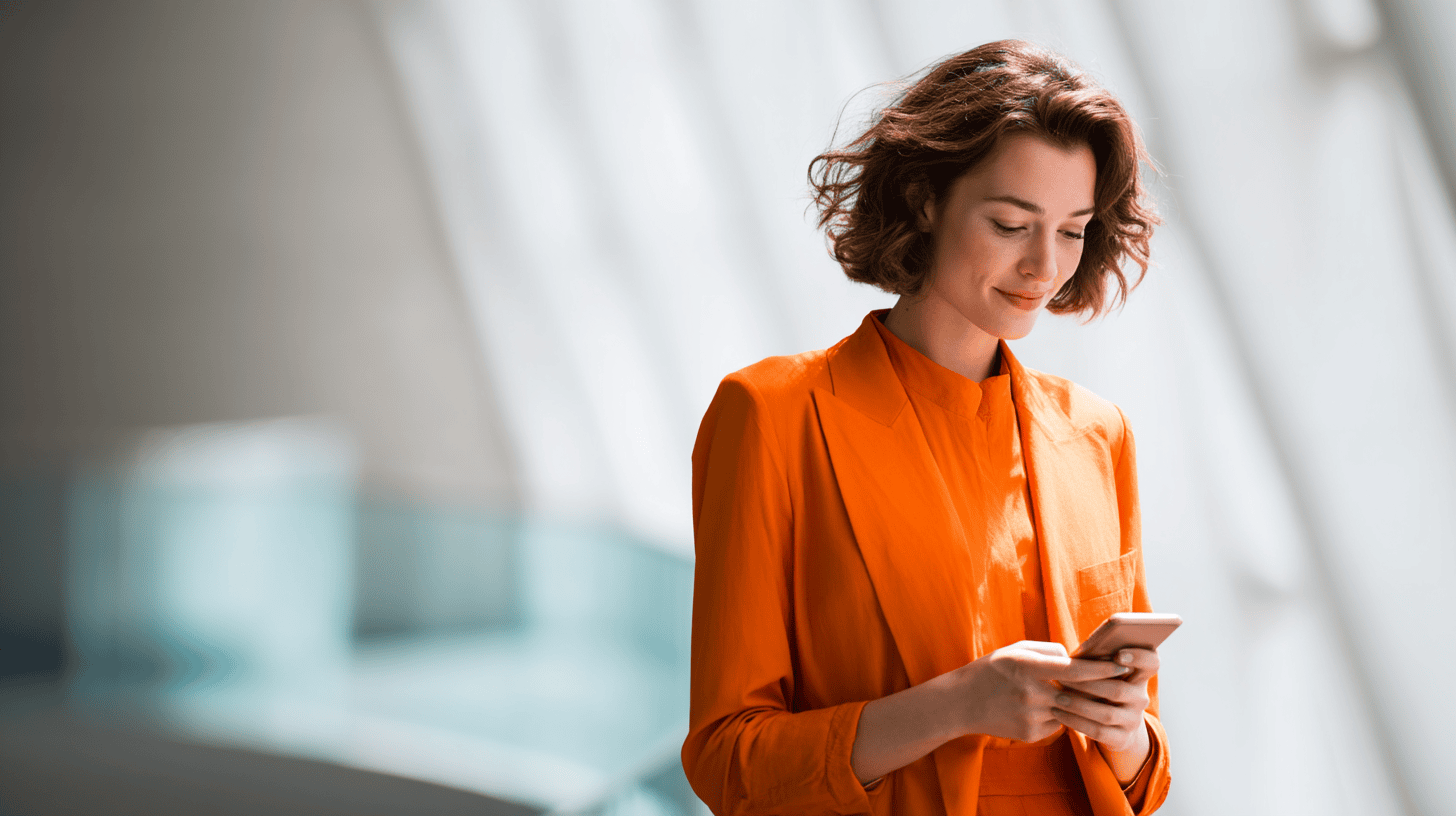 A woman with short, wavy hair wearing an orange suit is standing indoors, looking down at her smartphone. Soft natural light bathes the space, highlighting her focused expression and the modern architectural background.