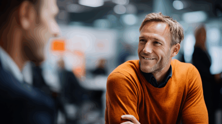 A man sitting in an office environment, wearing an orange sweater over a dark shirt, smiles while engaging in conversation. The background features blurred figures and light, suggesting a busy workplace atmosphere. The image conveys a friendly and professional interaction.