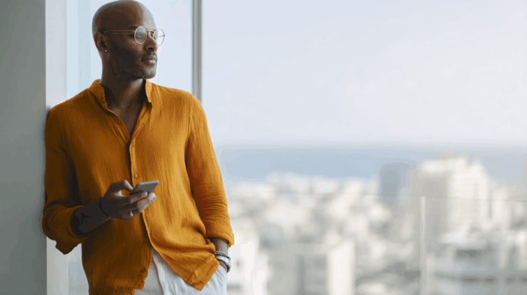 A man with a shaved head and glasses leans against a wall, holding a smartphone. He wears an orange button-up shirt and watches a cityscape with blurred buildings and a sea view in the background, creating a relaxed, contemplative atmosphere.
