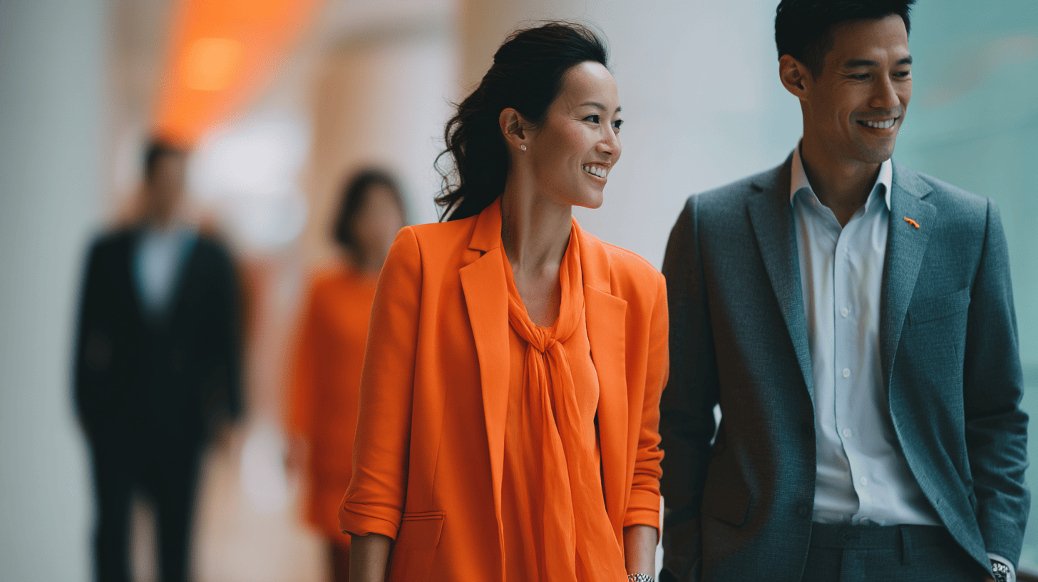 A woman in a vibrant orange blazer and blouse smiles while walking alongside a man in a gray suit and light blue shirt. In the background, two other individuals dressed in dark suits are walking. The setting features soft, warm lighting, creating a professional yet friendly atmosphere.