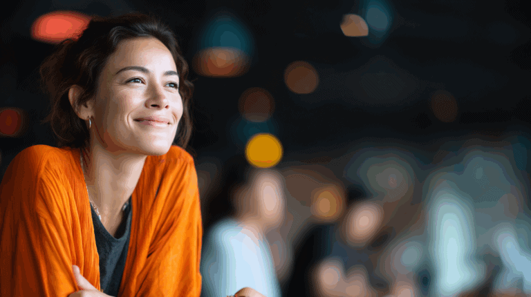 Smiling woman with curly hair wearing an orange cardigan, leaning on a table in a softly lit environment. The background includes blurred figures and warm, ambient light from hanging fixtures, creating a cozy atmosphere.