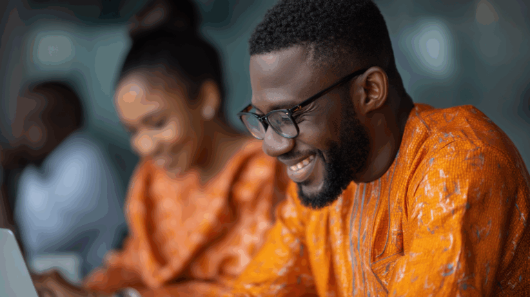 A man in glasses smiles while working on a laptop, wearing an orange traditional outfit. In the blurred background, a woman dressed similarly appears focused on her own laptop, creating a collaborative atmosphere. The setting suggests a modern workspace.
