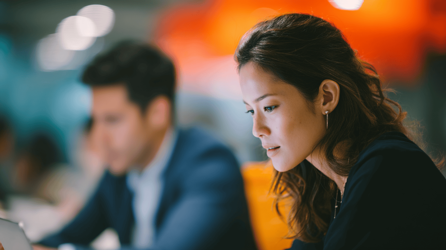 A woman with long, wavy hair is focused on a tablet, appearing thoughtful and engaged. In the background, a man is partially visible, also working on a device, in a modern workspace with blurred lighting and colors. The ambiance suggests a collaborative and creative environment.