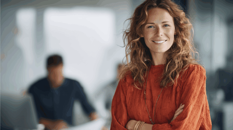 A smiling woman with wavy brown hair wearing an orange blouse stands confidently with her arms crossed. She is in a modern office setting with blurred colleagues in the background, suggesting a collaborative environment. The atmosphere is bright and professional, highlighting a positive workplace.