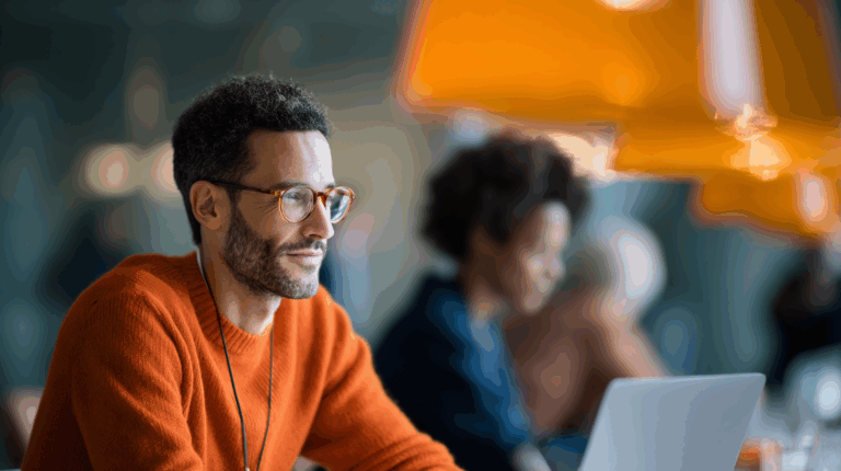A man with curly hair and glasses smiles while focused on his laptop in a bright workspace. He wears an orange sweater and is seated at a table with blurred figures in the background, including a woman with curly hair. Orange pendant lights hang above the setting, creating a warm atmosphere.