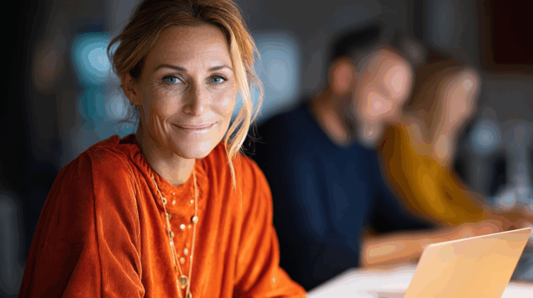 A smiling woman with long, wavy hair sits at a table, wearing an orange shirt and a necklace. She looks directly at the camera, exuding warmth and approachability. In the background, two blurred individuals are engaged in work on computers, suggesting a collaborative environment.