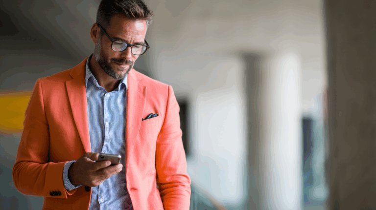A stylish man in an orange blazer and a striped shirt is looking at his smartphone. He has glasses and a well-groomed beard, with a focused expression. The background features a modern, softly blurred environment, highlighting the man as the main subject.
