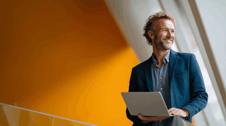 A smiling man in a blue blazer stands holding a laptop, gazing thoughtfully while positioned near large windows. The backdrop features a vibrant orange wall, enhancing the modern and professional ambiance of the setting.