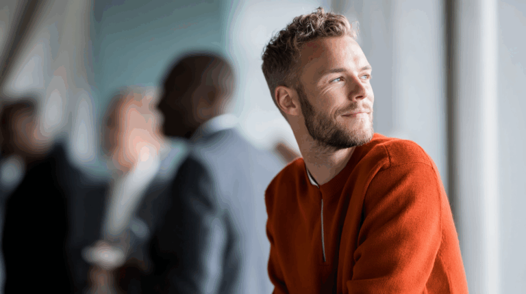 A young man with a beard and curly hair gazes thoughtfully while wearing an orange sweater. He is seated near a window, with soft natural light illuminating his face. In the background, blurred figures suggest a casual gathering or event, adding a lively atmosphere to the scene.