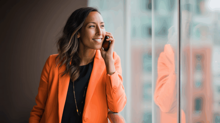 A smiling woman in an orange blazer is talking on her cellphone while standing next to a large window with a blurred urban background. She has wavy hair and is wearing a black top with a gold necklace, conveying a professional and engaged demeanor.