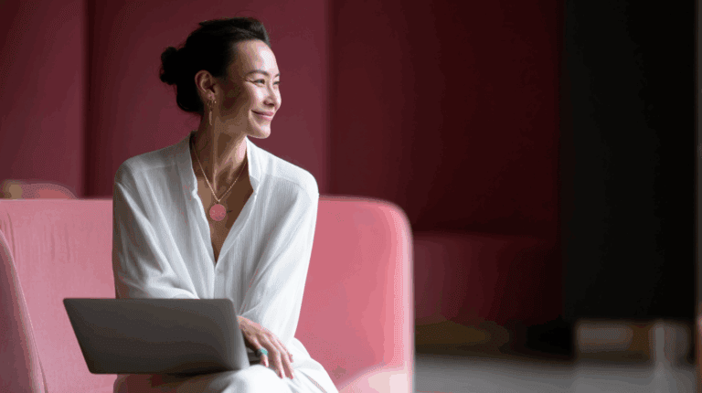 A smiling woman in a white shirt sits on a pink sofa, looking thoughtfully to her right while using a laptop on her lap. She wears gold earrings and a pendant necklace, with her hair styled in a bun. The background features soft red tones, creating a warm and inviting atmosphere.