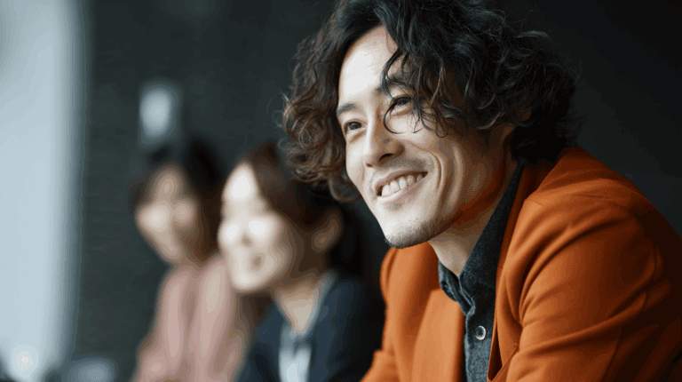 A man with curly hair and a warm smile is seen in the foreground, wearing an orange blazer over a dark shirt. Behind him, two out-of-focus individuals are slightly visible, contributing to a casual, friendly atmosphere. The setting appears modern and stylish.