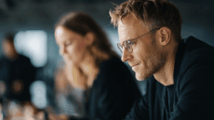 A focused man with glasses sits at a table, deep in thought. In the background, a woman with wavy hair partially visible is also engaged in a conversation. The setting appears to be a modern office or café, with a blurred, soft-focus ambiance that suggests a professional or creative meeting atmosphere.