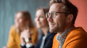 A focused man with glasses and a well-groomed beard sits at a table, wearing an orange blazer and a striped shirt. In the background, two blurred individuals are visible, one in an orange top. The setting suggests a professional or creative environment, with a soft-focus wall adding to the ambiance.