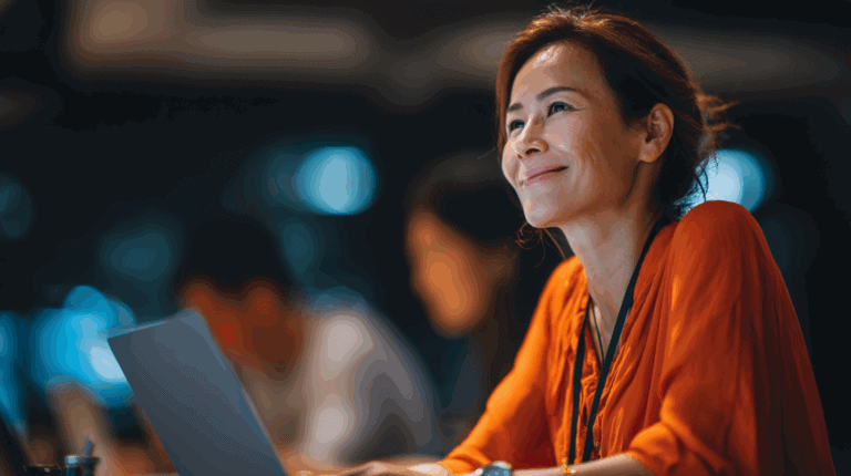 A woman with a warm smile is seated at a desk, focused on her laptop while wearing an orange top. The background is softly blurred, hinting at a lively workspace with other individuals in the distance. The overall atmosphere conveys a sense of engagement and positivity.