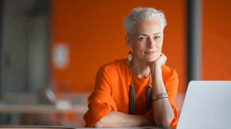 A confident older woman with short, silver hair smiles while resting her chin on her hand. She wears an orange blouse and statement earrings, seated at a desk with a laptop. The background features an orange wall, creating a warm and vibrant atmosphere.