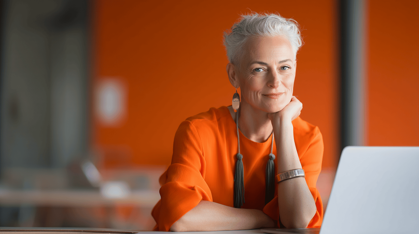 A confident older woman with short, silver hair smiles while resting her chin on her hand. She wears an orange blouse and statement earrings, seated at a desk with a laptop. The background features an orange wall, creating a warm and vibrant atmosphere.