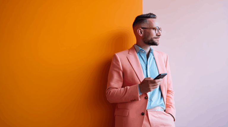 A man in a stylish pink suit and light blue shirt stands against a vibrant orange wall, looking thoughtfully to the side while holding a smartphone in his hand. The background transitions to a soft pink on his right side, creating a modern and colorful atmosphere.