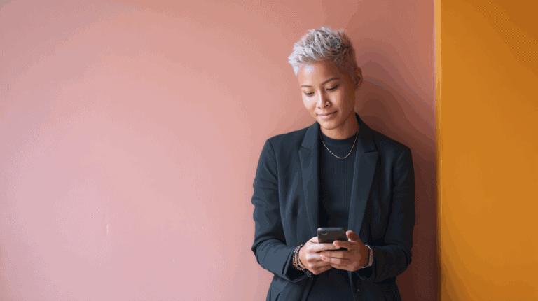 A person with short, stylish silver hair is standing against a pink wall, wearing a black blazer and a black top. They are holding a smartphone and looking at the screen with a slight smile, exuding a relaxed and confident demeanor. The background also features a contrasting orange corner.