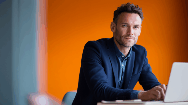 A professional man with short brown hair and a beard sits at a desk, looking at the camera with a warm smile. He wears a dark blazer over a blue shirt, with a laptop in front of him, and an orange wall serves as the background, creating a vibrant and inviting atmosphere.