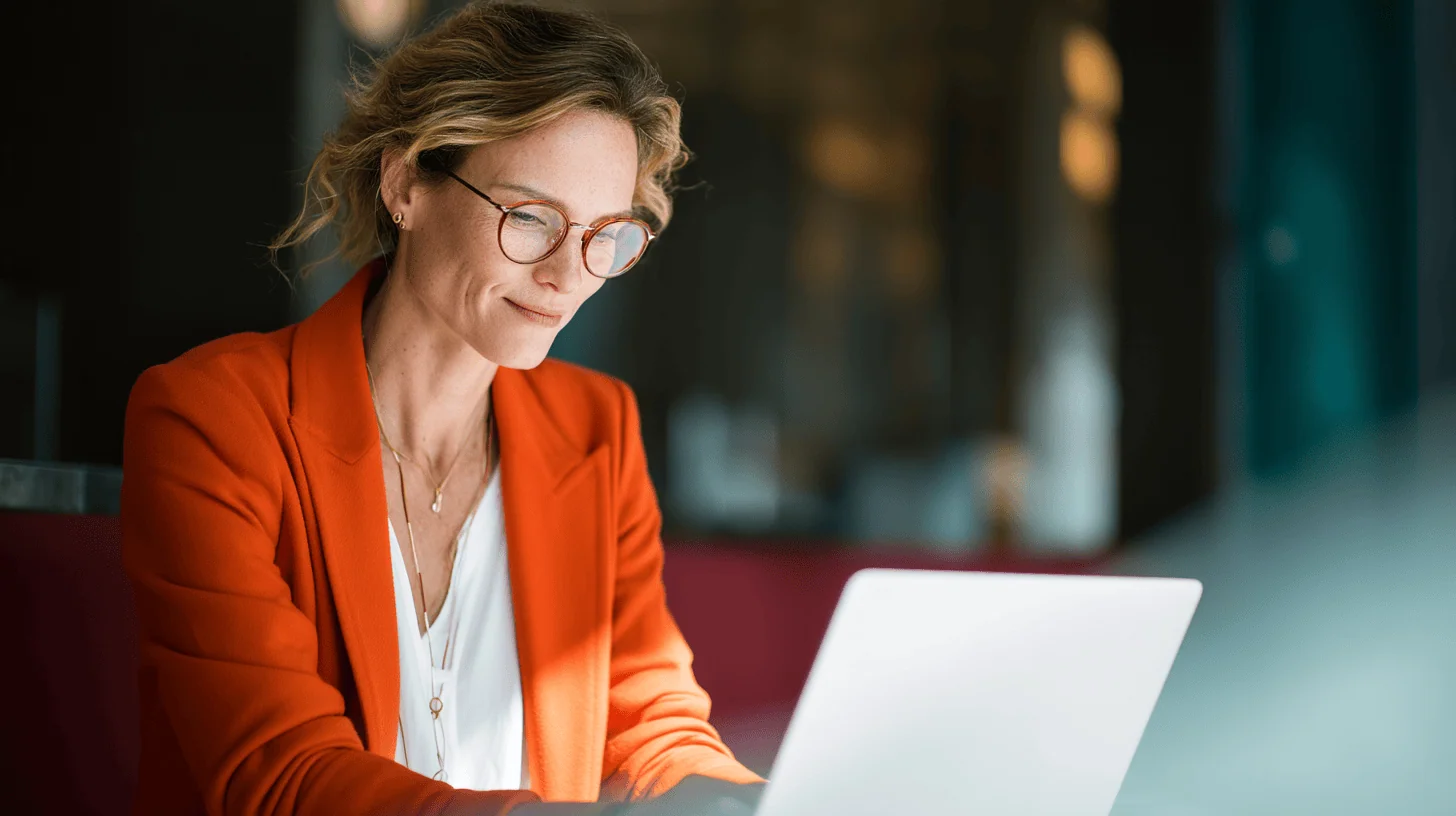 A woman in an orange blazer types on a laptop, focused, in a modern workspace with soft lighting.