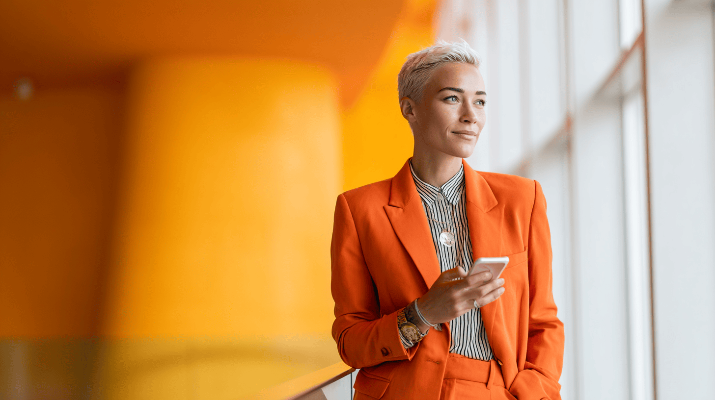 A woman in an orange suit stands by a window, holding a smartphone, with a bright orange background.