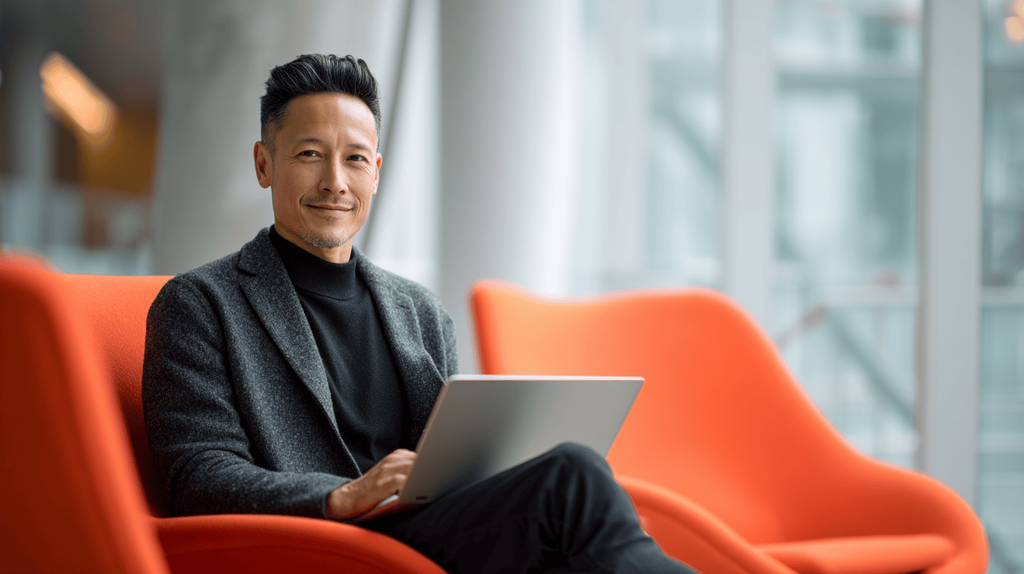 A man sitting in an orange chair with a laptop, smiling and wearing a gray jacket and black turtleneck. Bright, modern setting.