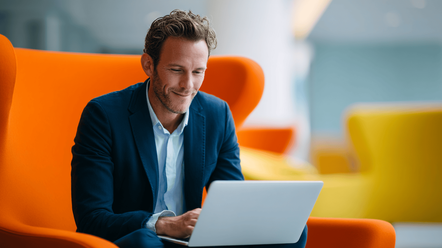 Man in a navy blazer sitting in an orange chair, using a laptop with colorful chairs in the background.