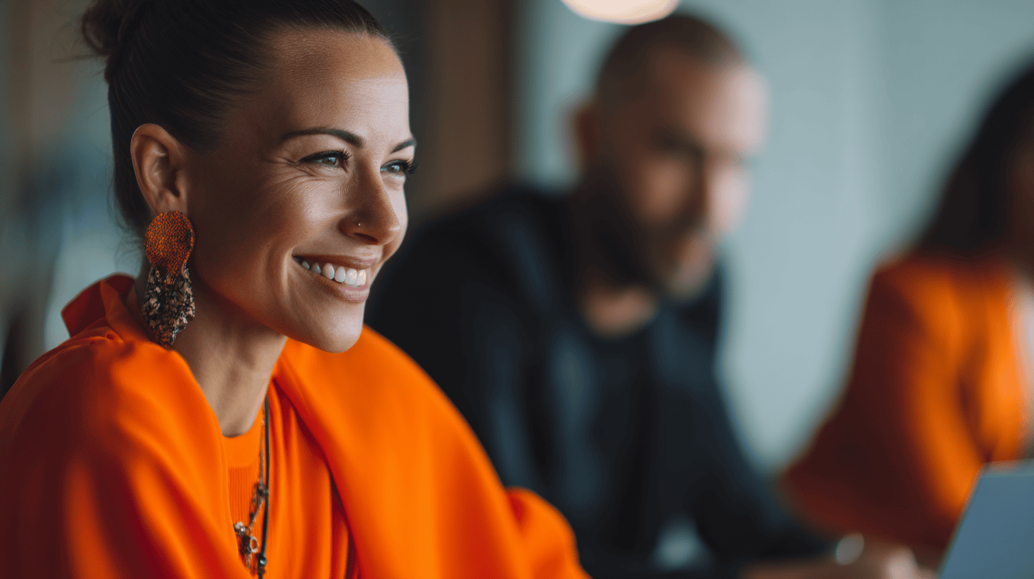 A smiling woman in an orange top and statement earrings, seated with two blurred colleagues in a meeting setting.