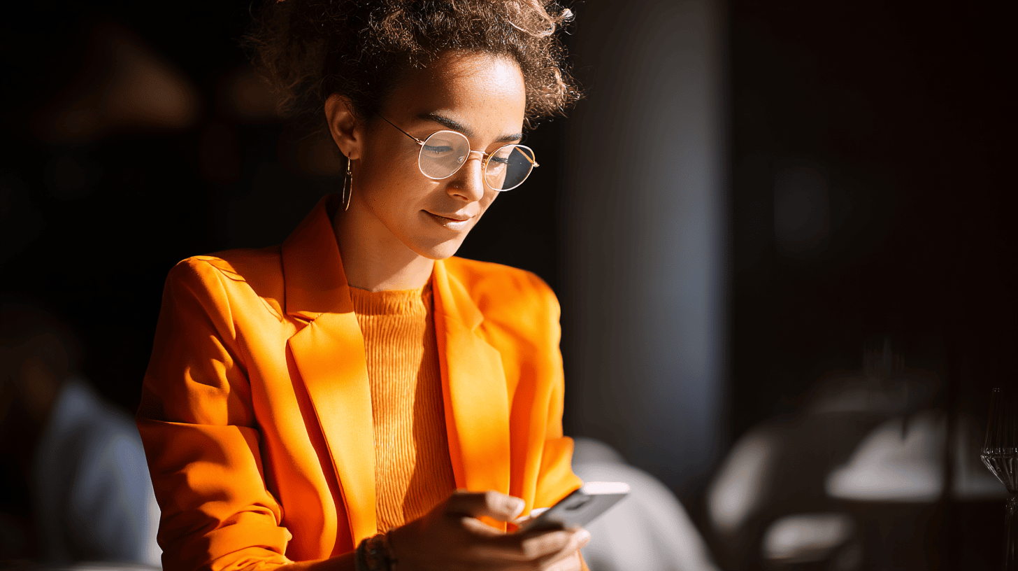 A woman in an orange blazer uses her smartphone in a softly lit indoor setting.