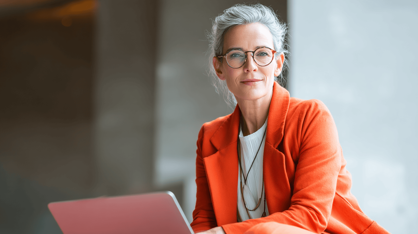 A confident woman in an orange blazer sits with a laptop, looking directly at the camera in a well-lit, modern setting.