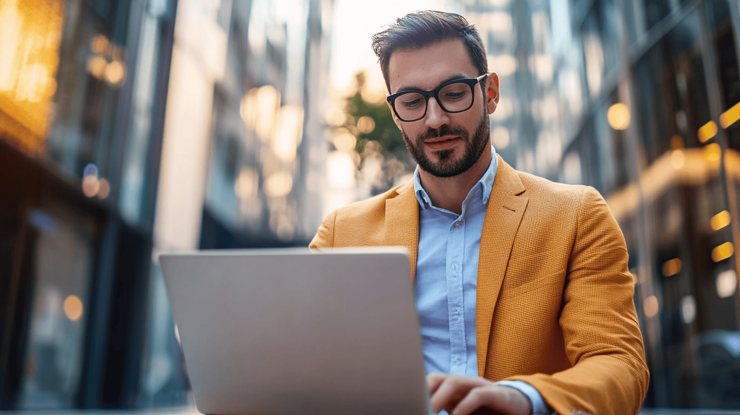 A man in an orange blazer uses a laptop outdoors, surrounded by modern buildings and greenery.
