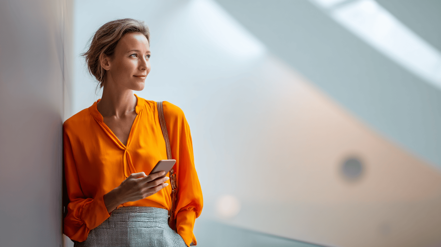 A woman in an orange blouse stands by a wall, looking at her smartphone in a bright, modern setting.
