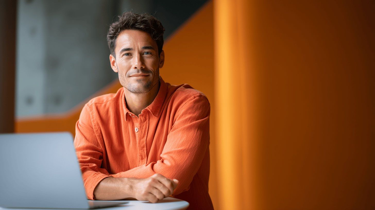 A smiling man in an orange shirt sits at a table with a laptop, against a backdrop of orange and gray geometric shapes.