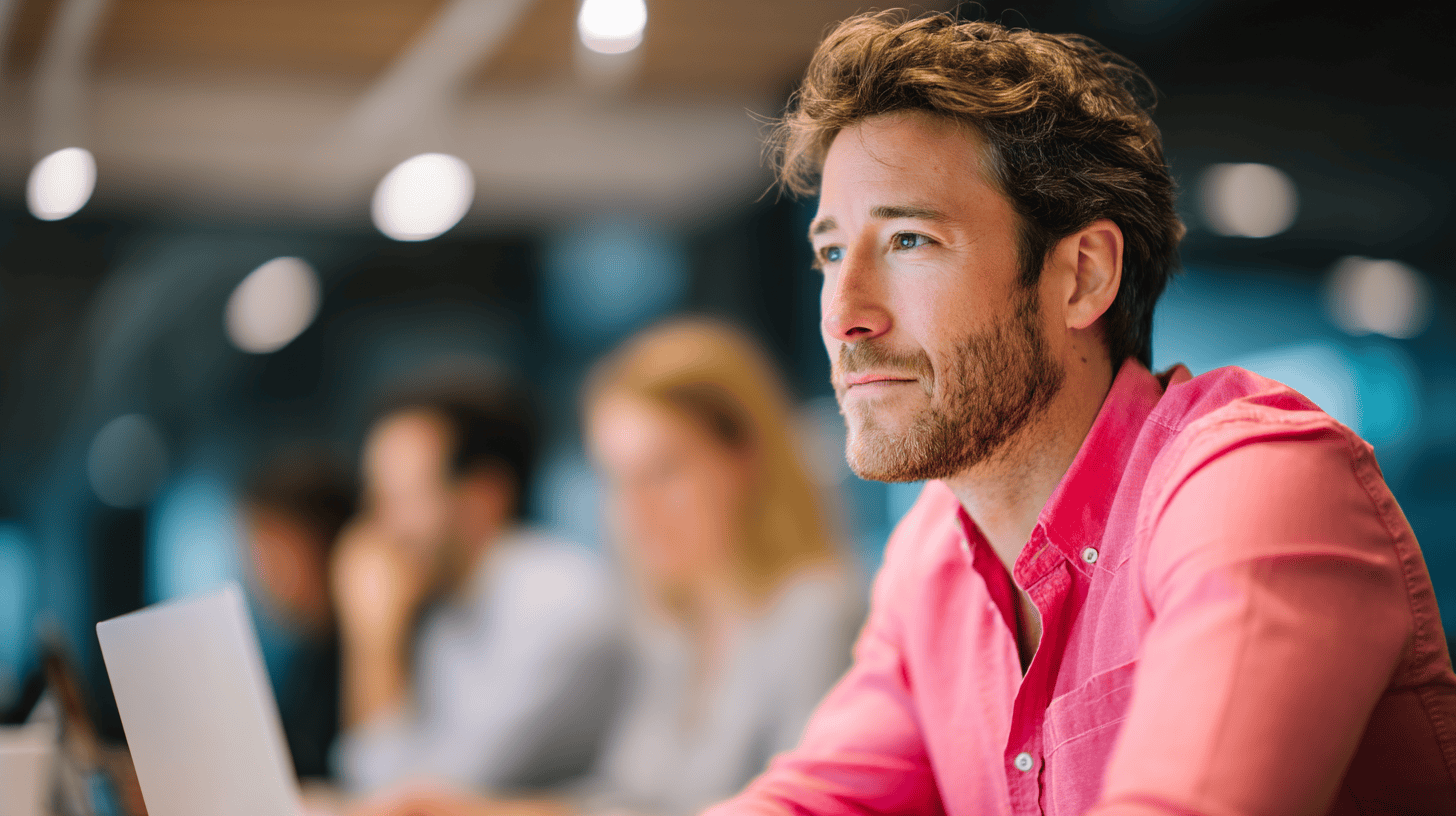 Man in a pink shirt looking thoughtfully to the side in a modern workspace; laptop and blurred colleagues in background.