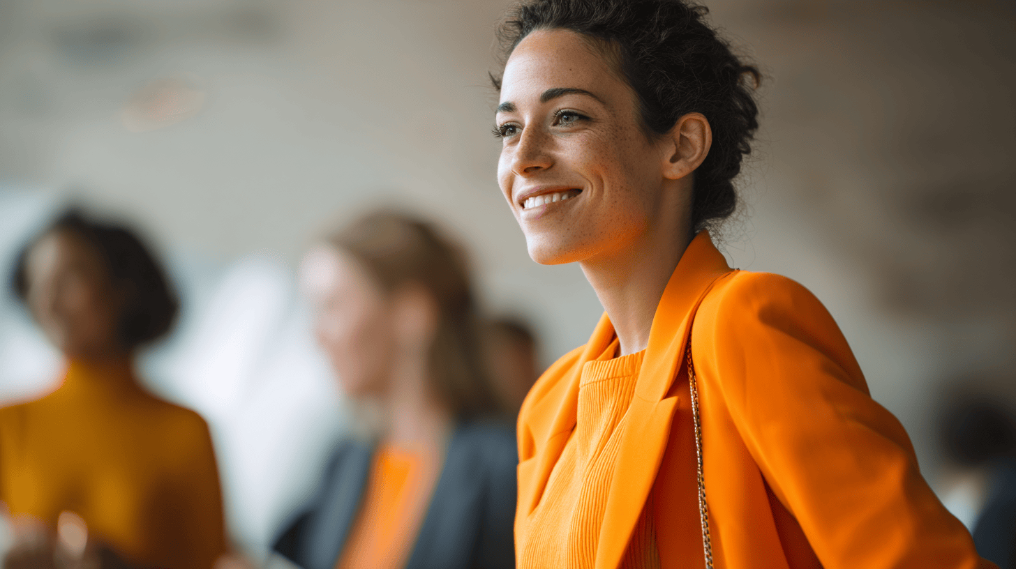 A woman in an orange jacket smiles, engaging with blurred figures in the background at a professional event.