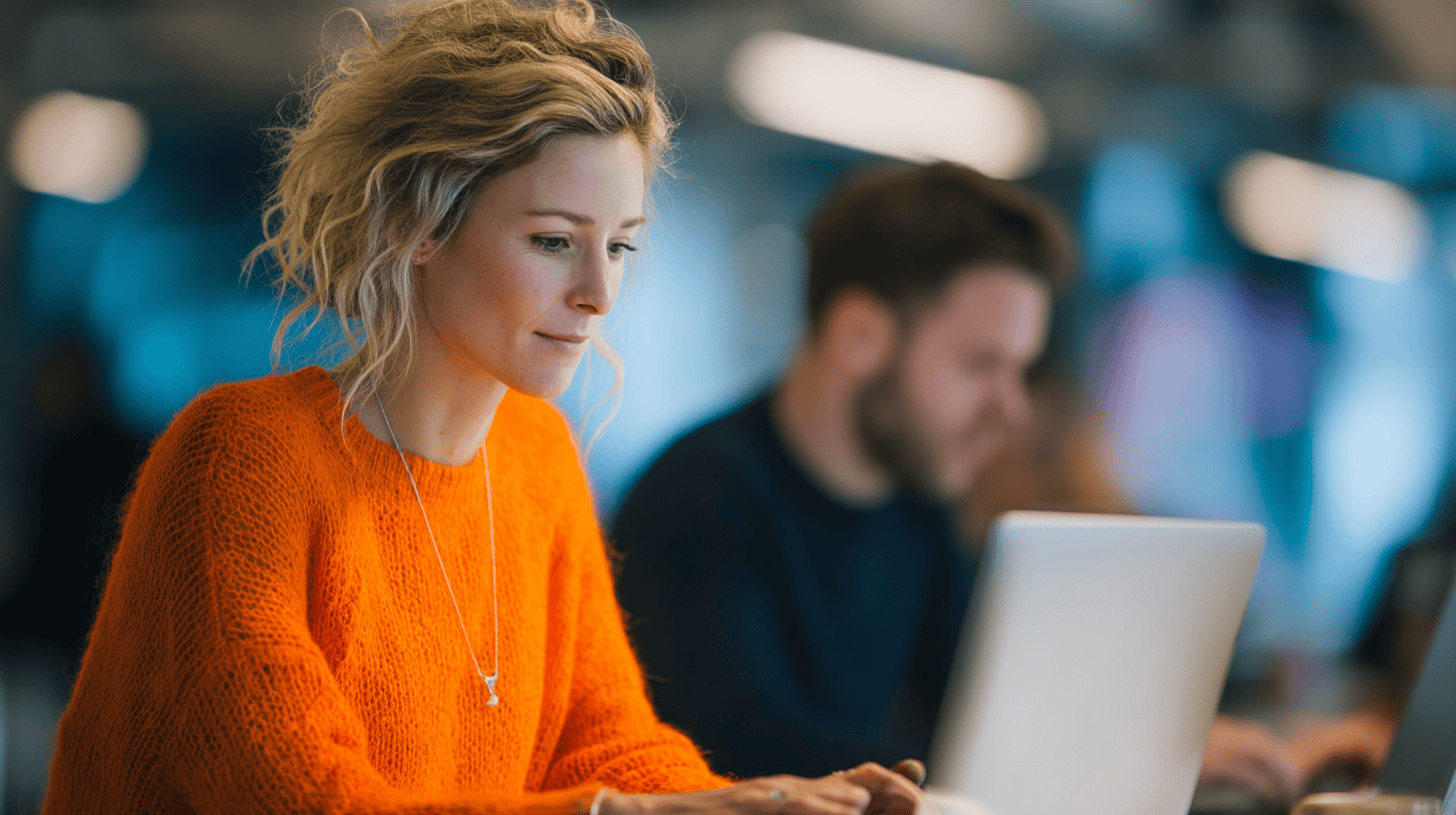 A woman in an orange sweater focuses on her laptop in a modern workspace, with a man working in the background.