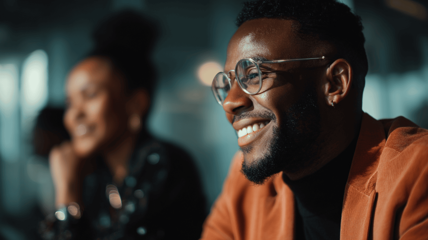 A smiling man in glasses and an orange jacket, with a blurred woman in the background, highlighted by natural light.