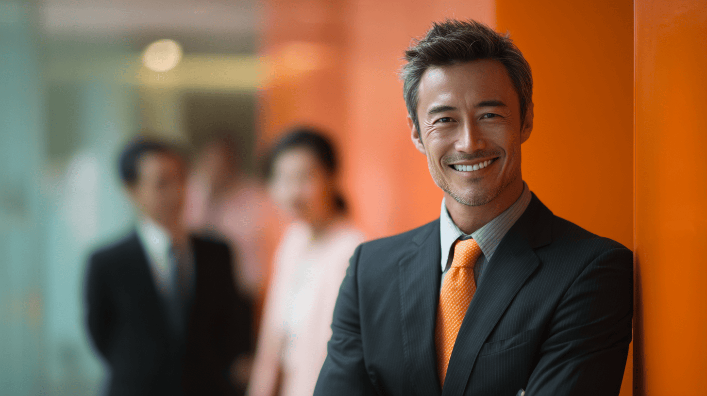 Smiling man in a suit with an orange tie stands against an orange wall, with blurred colleagues in the background.