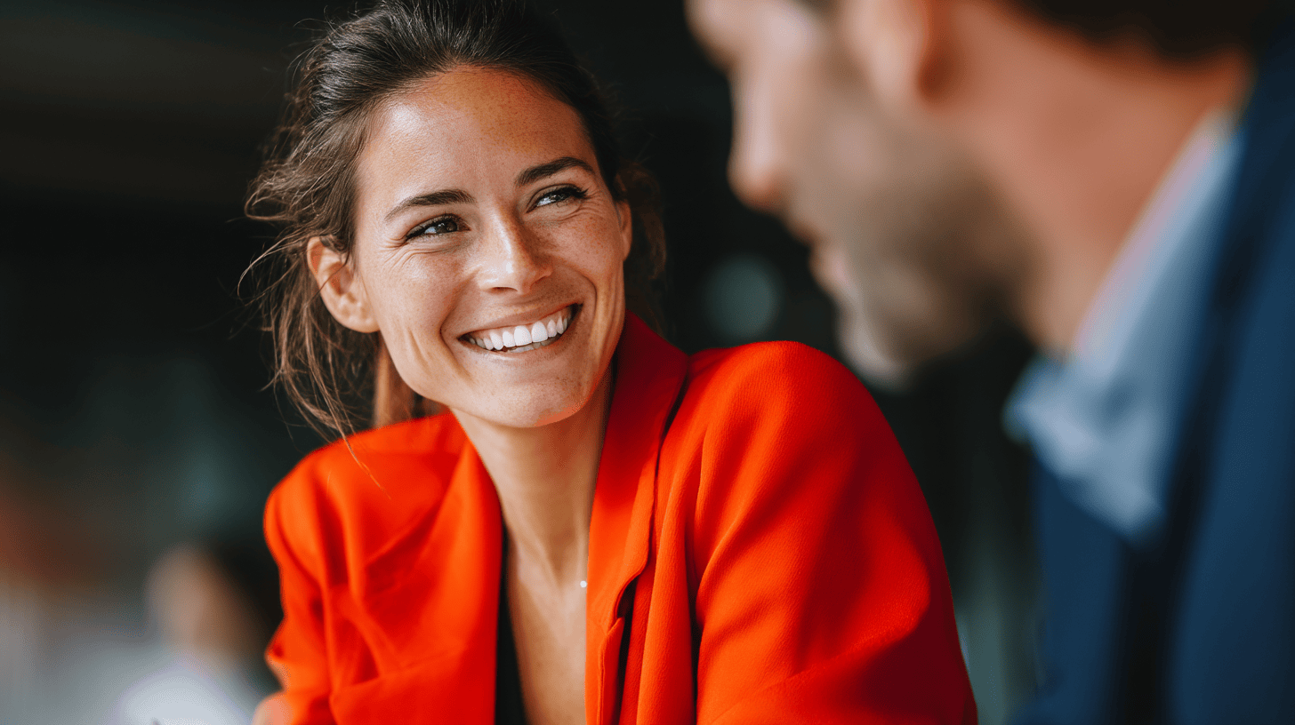 A woman in a bright orange blazer smiles warmly at a blurred man in a professional setting.