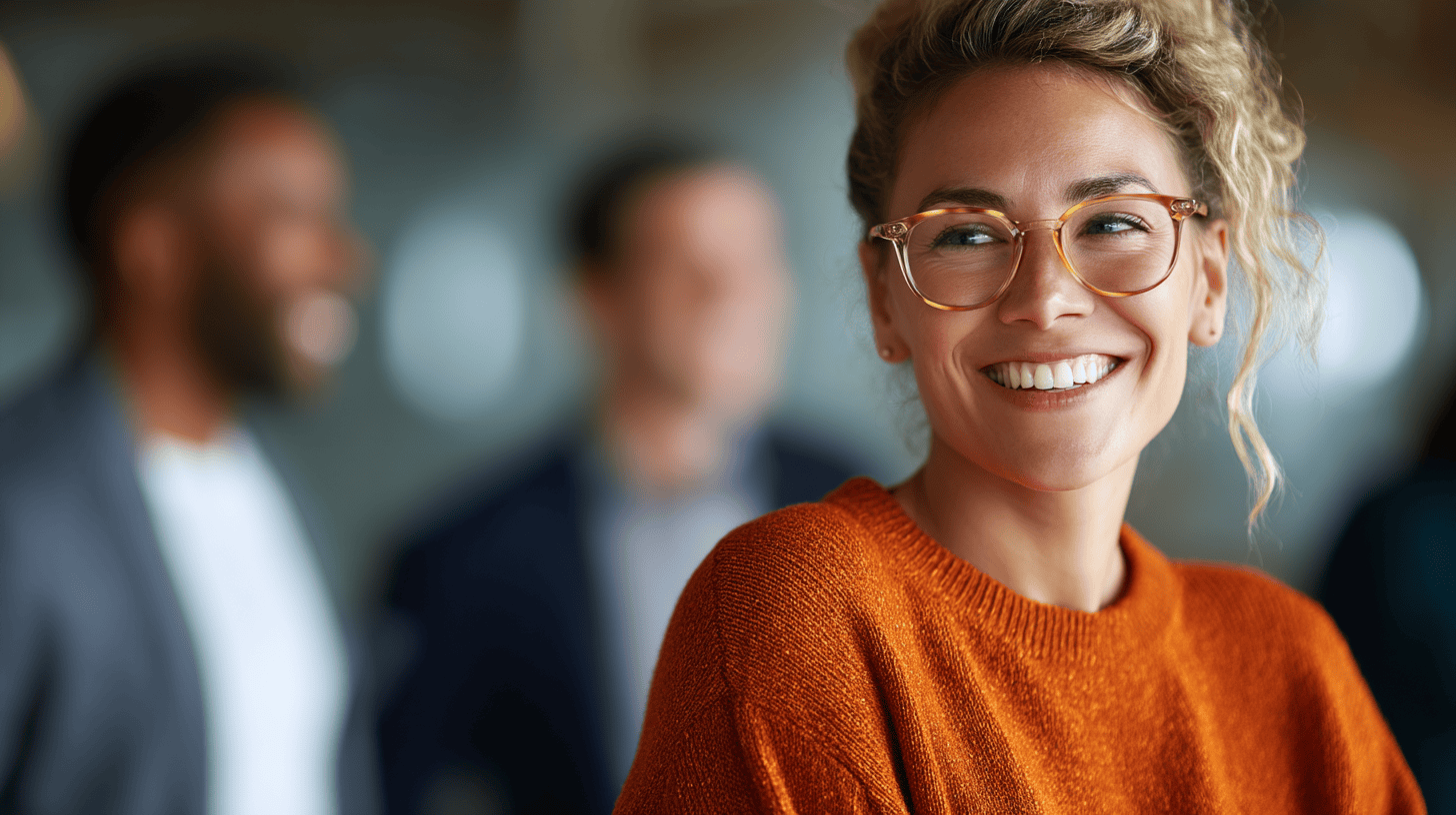 Smiling woman in an orange sweater and glasses, with blurred colleagues in the background.
