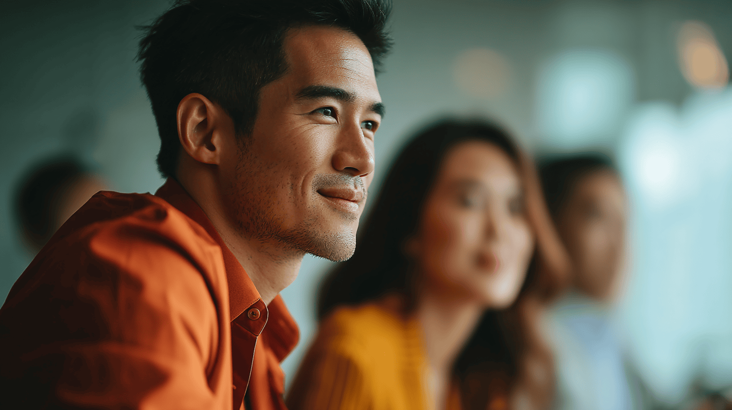 A smiling man in an orange shirt sits in focus, with two blurred people in the background during a meeting.